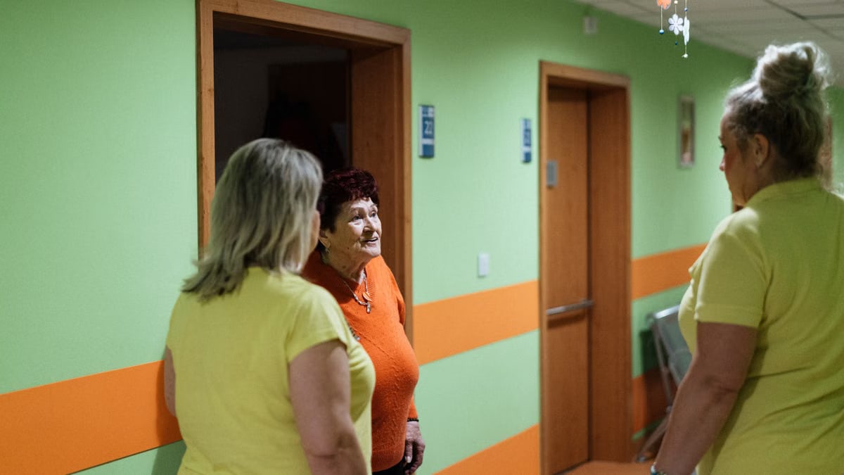 Senior woman engages in conversation with caregivers in a facility corridor.